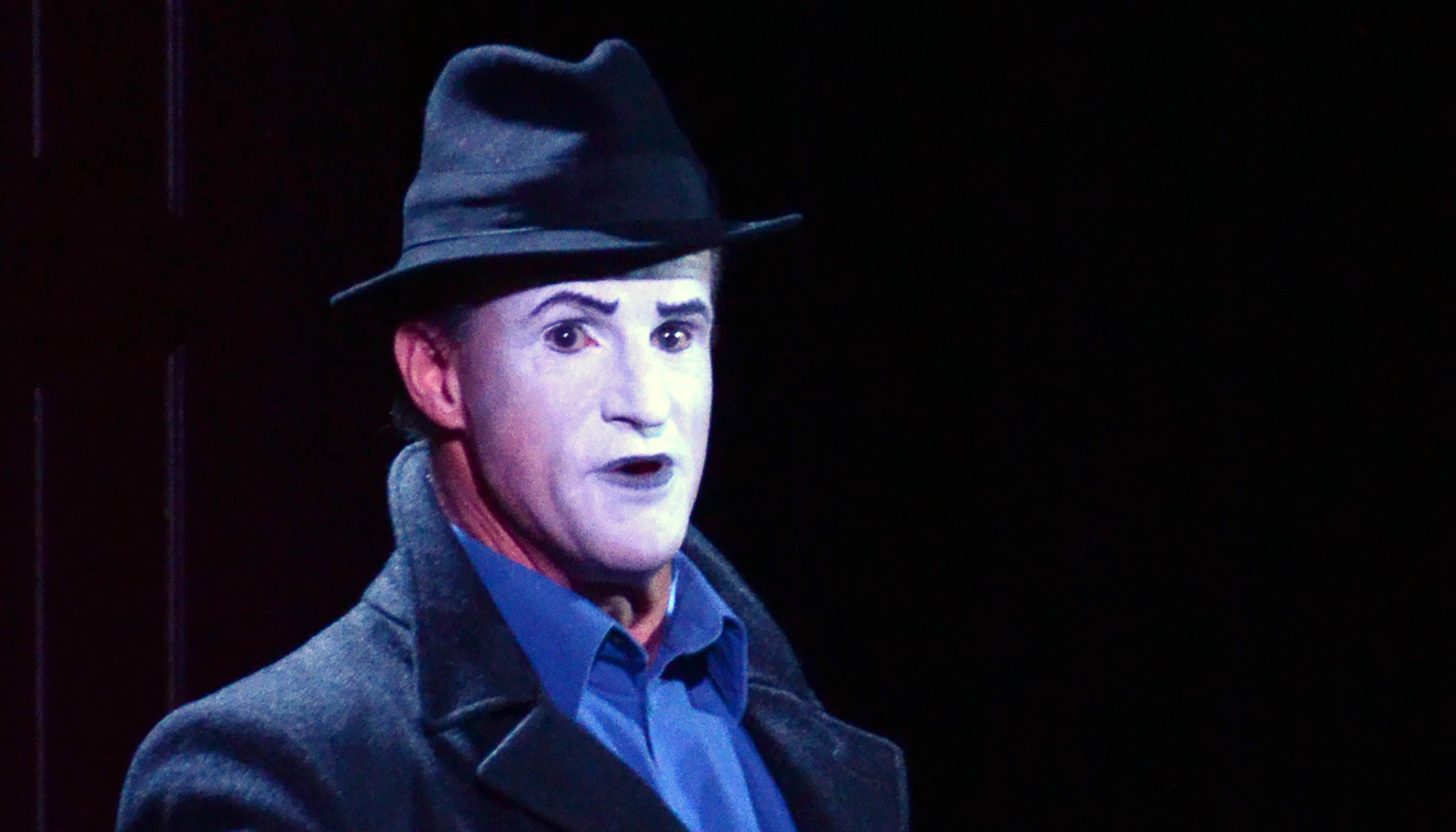 Lawton onstage, wearing intense makeup (white paint, arched black eyebrows), black overcoat, blue collared shirt, and black fedora.